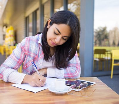 Focused young woman making notes in outdoor cafe. Pretty lady wearing casual clothes and sitting at table with chairs and building in background. Education concept. Front view.