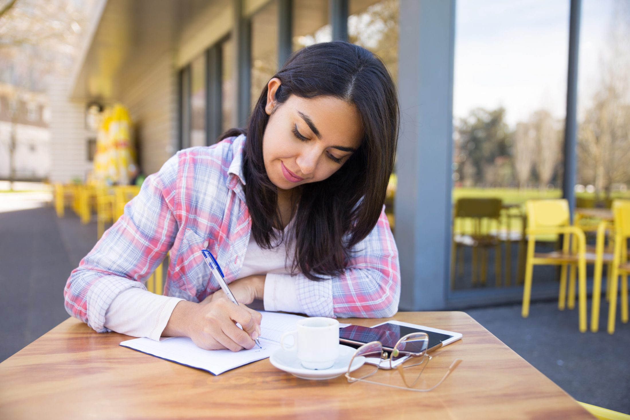 Focused young woman making notes in outdoor cafe. Pretty lady wearing casual clothes and sitting at table with chairs and building in background. Education concept. Front view.