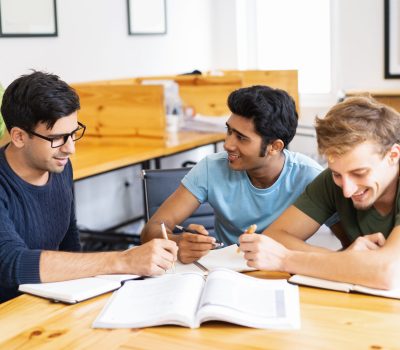 Three students studying and doing homework together. Young men talking, writing and sitting at desk in classroom or library. Education concept.