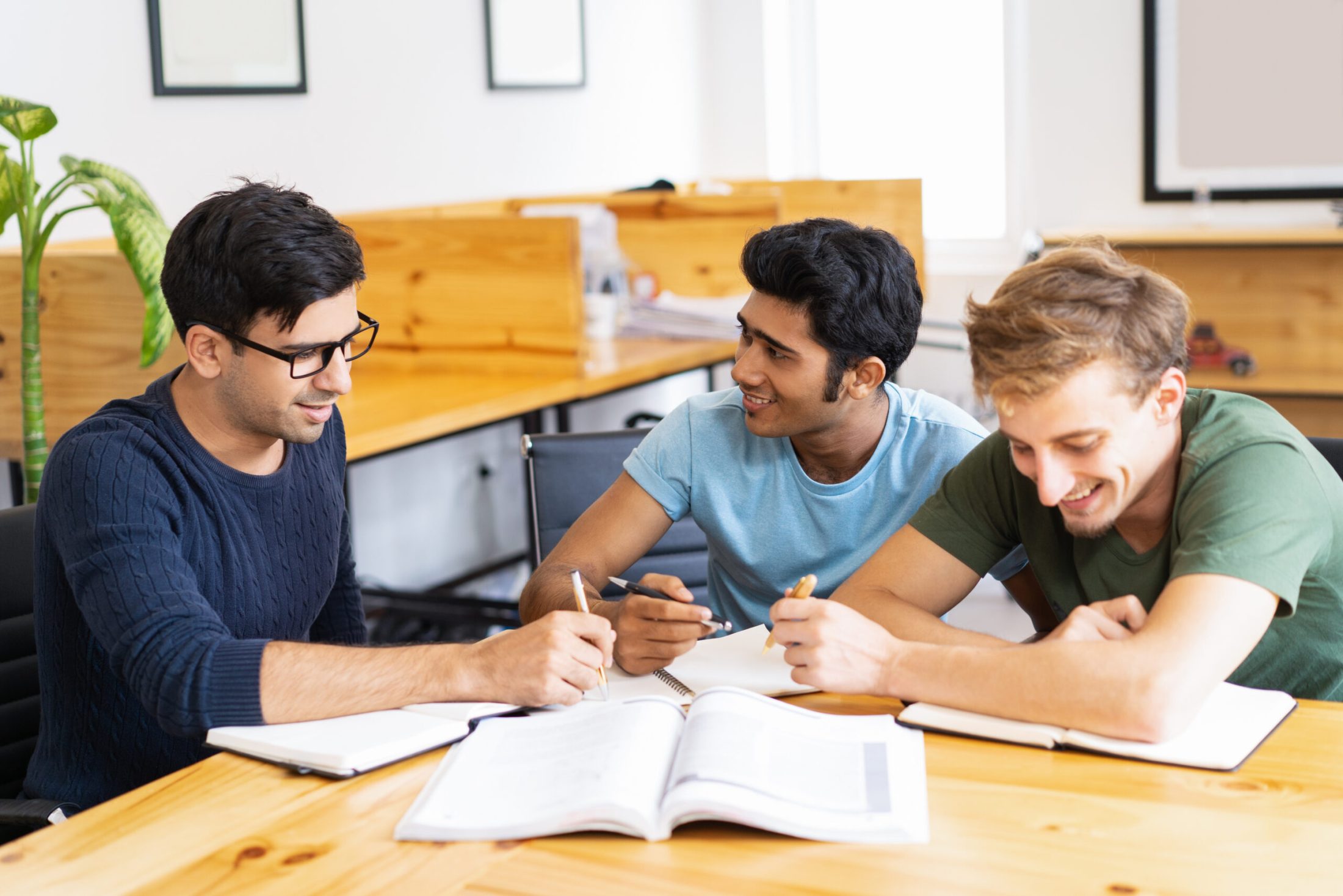 Three students studying and doing homework together. Young men talking, writing and sitting at desk in classroom or library. Education concept.